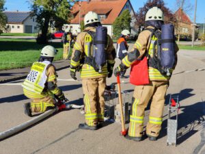 zwei Feuerwehrleute unter Atemschutz stehen bereit für ihren Einsatz. Ein weiterer kniet auf der Strasse und bedient den Wasserverteiler. Im Hintergrund sind weitere Feuerwehrleute erkennbar.