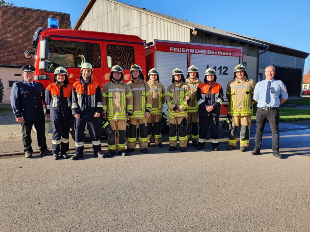 die acht Prüflinge stehen zusammen mit drei Prüfern in einer lockeren Linie vor dem Feuerwehrfahrzeug. Sie freuen sich über die bestandene Prüfung.