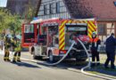 der Maschinist steht hinter dem Feuerwehrfahrzeug und bedient die Pumpe, während sich ein Trupp neben dem Fahrzeug mit Atemschutz ausrüstet.