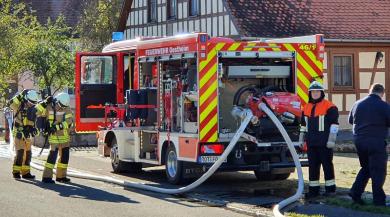 der Maschinist steht hinter dem Feuerwehrfahrzeug und bedient die Pumpe, während sich ein Trupp neben dem Fahrzeug mit Atemschutz ausrüstet.